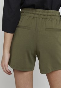 Olivgröna shorts med elastisk midja, som har två bakfickor och en mjuk, slät textur. Platt sydd fåll.