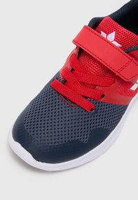 Červená a námořnická atletická obuv s prodyšným vrškem, suchým zipem a tkaničkami. Obsahuje bílou podrážku a texturované akcenty na špičce a patě.