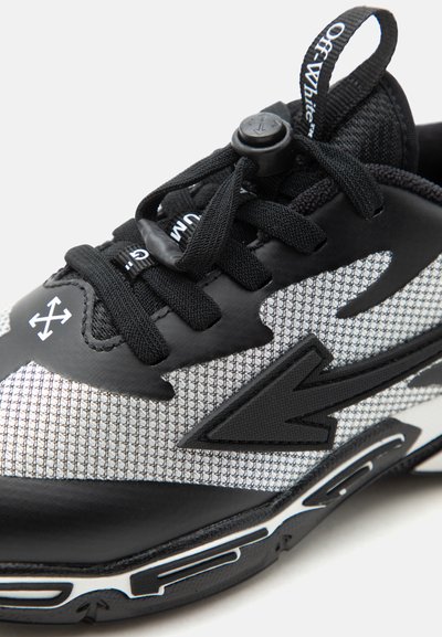 Κοντινό πλάνο ασπρόμαυρου αθλητικού παπουτσιού με υφασμένο διχτυωτό υλικό, μαύρα κορδόνια με κούμπωμα, λογότυπο βέλους και ετικέτα τράβηγματος με την επωνυμία "Off-White".