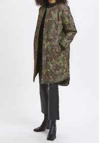 Kurtka pikowana w kamuflażowym wzorze w odcieniach zieleni i brązu, z okrągłym dekoltem, otwartym przodem i kieszeniami po bokach. Stylizowana z czarnymi spodniami i botkami.