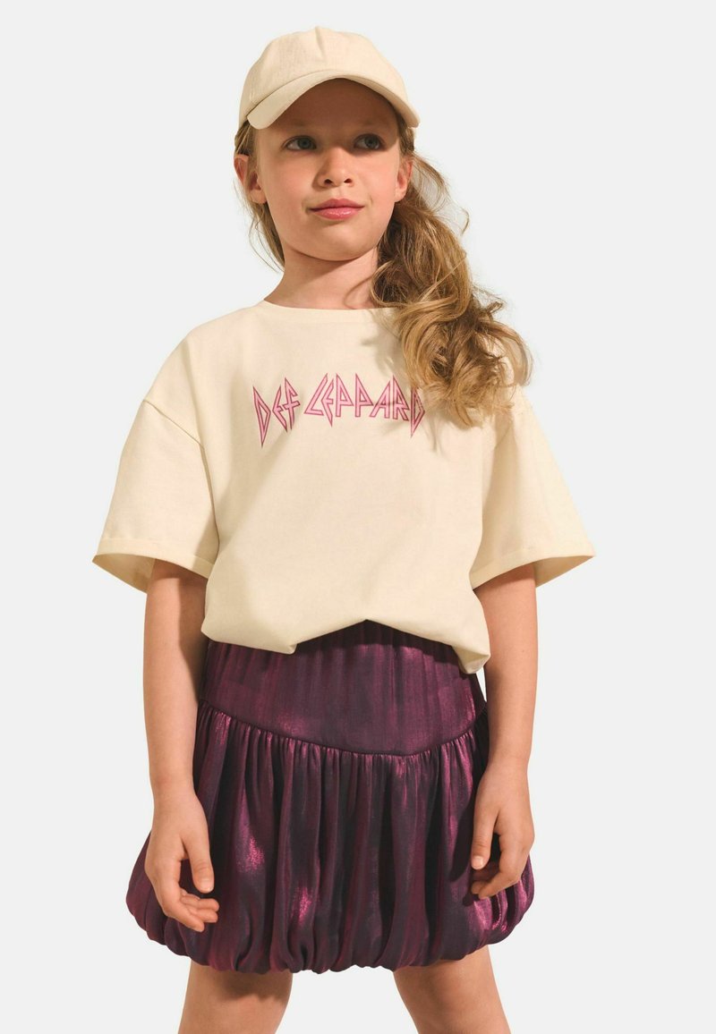 T-shirt crème à manches courtes avec un imprimé "Def Leppard" en rose, associé à une jupe plissée métallique violette. Casquette portée sur la tête.