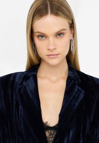 Blazer de terciopelo azul marino con solapas de muesca, combinado con una camisola de encaje negro. La modelo lleva unos pendientes largos y brillantes. Textura suave.