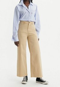 Chemise à rayures bleu clair et blanche avec des manches amples, associée à un pantalon large beige et des baskets montantes noires.