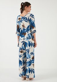 Completo a due pezzi composto da una camicia larga e pantaloni a gamba larga. Motivo di foglie blu e marrone su uno sfondo chiaro. Tessuto strutturato, maniche a tre quarti.