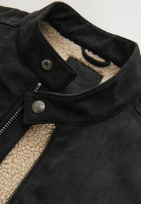 Sort ruskindsjakke med en fluffy beige fleece foring, trykknaplukning og diskrete syninger langs kraven og lynlåsen.