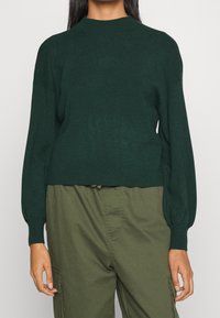 Tummanvihreä neulepusero pyöreällä pääntiellä, pitkillä hihoilla ja lyhyellä leikkauksella. Pehmeä pinta, jossa hieman kiiltoa, ja hihansuiden resorit.