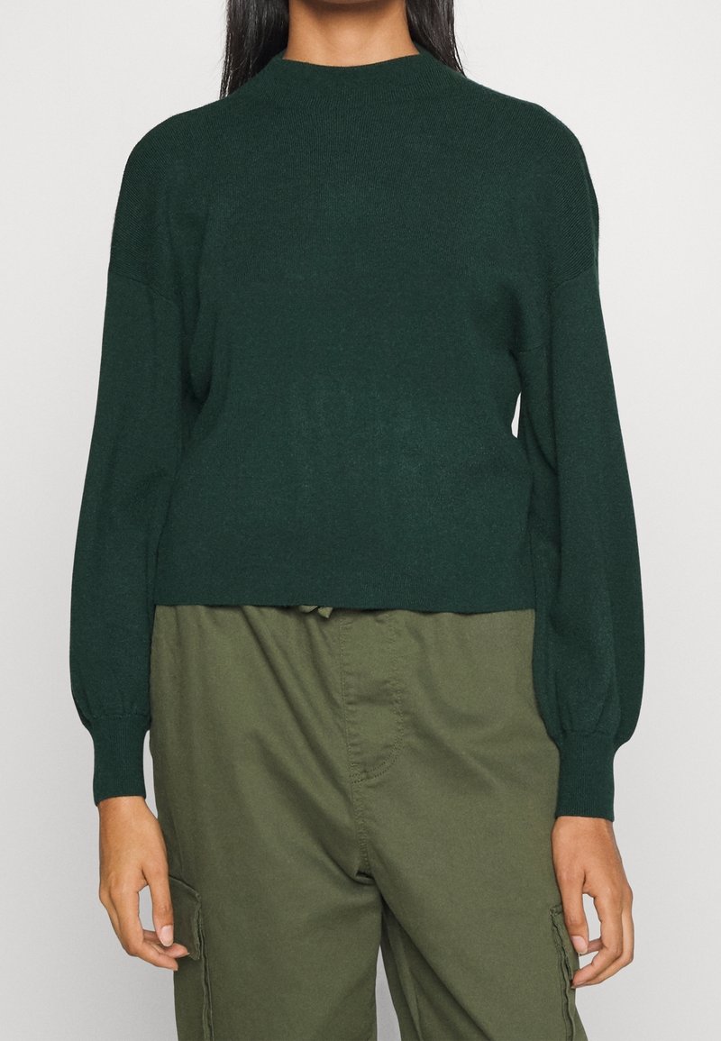 Tummanvihreä neulepusero pyöreällä pääntiellä, pitkillä hihoilla ja lyhyellä leikkauksella. Pehmeä pinta, jossa hieman kiiltoa, ja hihansuiden resorit.