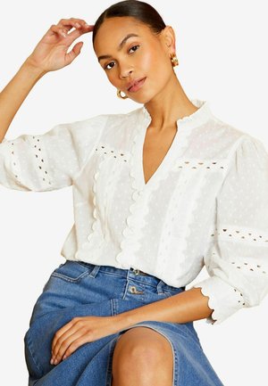 Žena nosi bielu blúzku s textúrou, ozdobenú vlnkovanými lemami a nafúknutými rukávmi, v kombinácii s modrou denimovou sukňou, pózuje s rukou pri vlasoch.