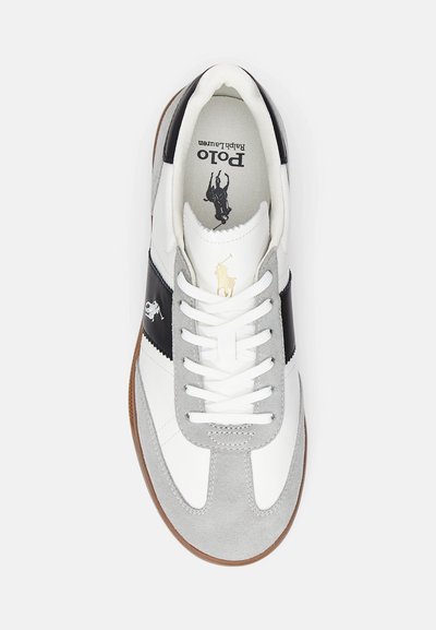 Adidași albi și gri cu vârful din suede gri texturat, dungi negre și un logo auriu. Prezintă șireturi albe și o talpă din cauciuc.