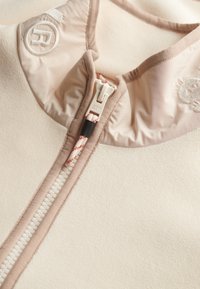 Capo in pile beige chiaro con colletto alto, dotato di una cerniera liscia e un cordoncino. Tessuto strutturato con dettagli di cuciture sottili.