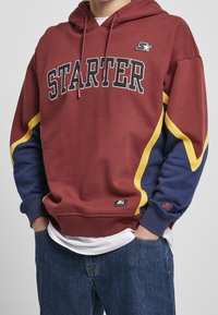 Sweat à capuche bordeaux avec logo "STARTER" noir, accents jaunes et bleus, et poignets côtelés. Comprend une poche avant et des cordons de serrage réglables.