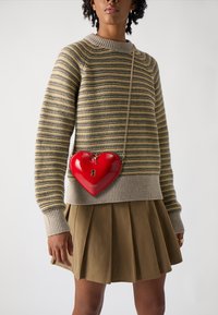 Red heart-shaped crossbody bag with a shiny finish, zipper closure, and keyhole detail, paired with a striped sweater and pleated skirt.