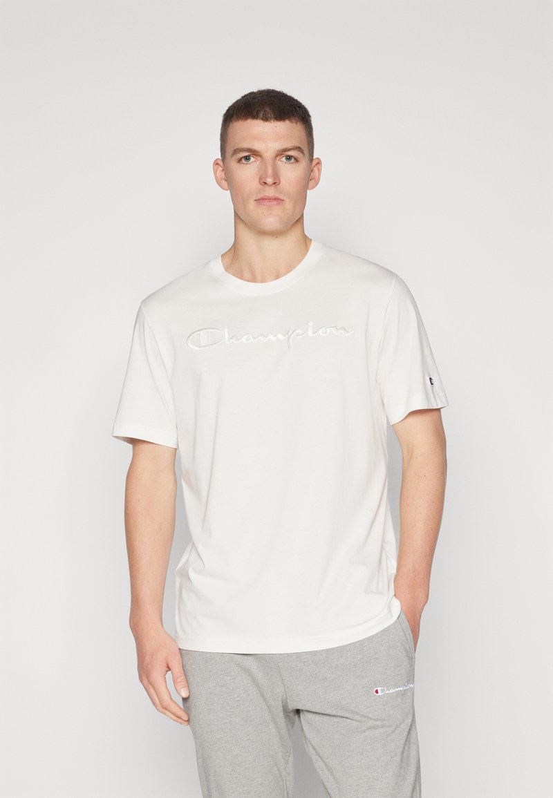 Weißes Baumwoll-T-Shirt mit gesticktem „Champion“-Logo in Hellgrau. Rundhalsausschnitt und kurze Ärmel, kombiniert mit grauen Sweatpants.