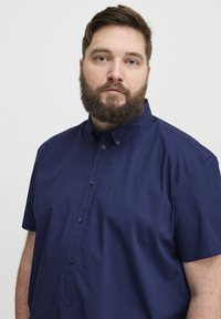Tamsiai mėlynas trumpomis rankovėmis mygtukų marškinis iš sklandaus medvilnės audinio, su klasikiniu apykakle ir subtilia siuvimo detale palei mygtukų eilę.