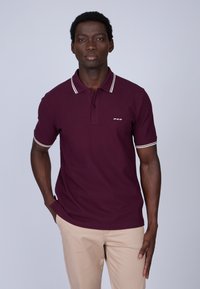 Polo maroon avec des bordures blanches au col et aux manches, présentant une patte de boutonnage à trois boutons et un petit logo sur la poitrine. Porté avec un pantalon beige.
