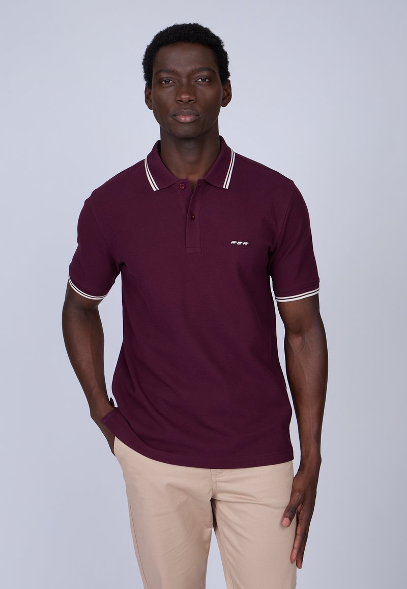 Polo maroon avec des bordures blanches au col et aux manches, présentant une patte de boutonnage à trois boutons et un petit logo sur la poitrine. Porté avec un pantalon beige.
