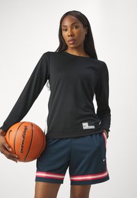 Musthave musta pikkade varrukatega spordisärk, millel on Nike silt, koos tumesiniste lühikesed püksidega, millel on roosad külje triibud. Modell hoiab korvpalli.