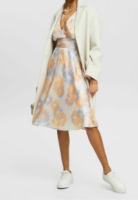 Robe florale avec des motifs orange et bleu, décolleté en V, silhouette en A, tissu en chiffon superposé, accompagnée d'un manteau léger et de baskets blanches.