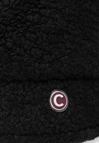 Fekete bolyhos sapka texturált felülettel. Jellemzője egy szürke kör alakú címke, amelyen egy bordó "C" látható. Az anyag puhanak és vastagnak tűnik.