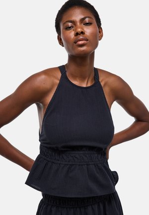 Jeune femme noire portant un top noir sans manches avec un peplum à la taille, posant avec les mains sur les hanches devant un fond uni.