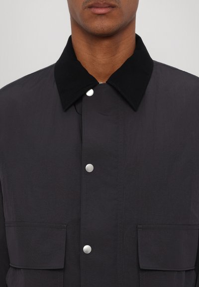 Fekete dzseki texturált anyaggal, patentgombos záródással és eltérő fekete gallérral. Elöl zsebek találhatóak a praktikum érdekében.