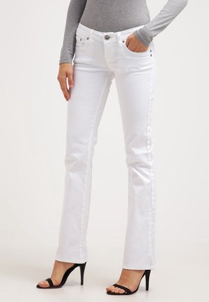 Femme portant un jean bootcut blanc, un haut gris à manches longues et des sandales noires à talons hauts et à bouts ouverts, debout devant un fond uni.
