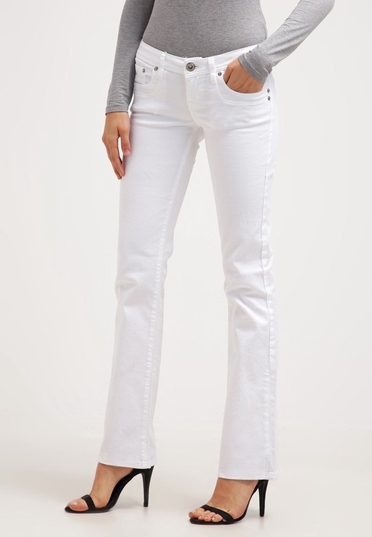 Femme portant un jean bootcut blanc, un haut gris à manches longues et des sandales noires à talons hauts et à bouts ouverts, debout devant un fond uni.