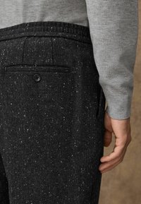 Dunkelgraue Fischgrät-Hosen mit einer gesprenkelten Textur, ausgestattet mit einer einzigen Gesäßtasche und einem Knopfverschluss, kombiniert mit einem hellgrauen Pullover.