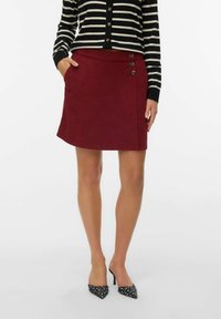 Femme portant une jupe bordeaux à boutons, un haut à rayures noires et blanches à manches longues, et des chaussures à talons hauts noires avec des pois blancs.
