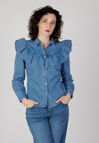 Camicia di denim con un ampio colletto arricciato, maniche lunghe e chiusura con bottoni. Indossata con pantaloni in denim abbinati, di un blu medio uniforme.