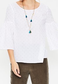 Blusa blanca con cuello redondo, tela texturizada y mangas abullonadas. Presenta pequeños patrones de puntos dorados y un collar decorativo con borlas azules.