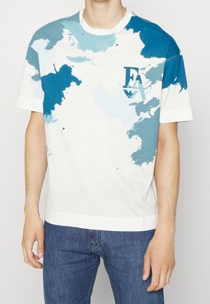 Mann trägt weißes T-Shirt mit blauem und türkisfarbenem abstraktem Farbspritz-Design und "FAY" Logo auf der Brust, kombiniert mit blauen Jeans.