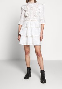 Valkoinen kerroksellinen mekko, jossa on pitsiyksityiskohtia, röyhelösateet ja korkea kaulus, yhdistettynä mustiin nauhakengät nilkkamittaisina.