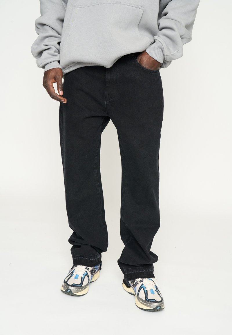 Pantalones de mezclilla negra con corte recto, combinados con una sudadera con capucha de color gris claro. Acentuados por zapatillas modernas con detalles en azul.