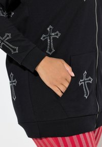 Felpa nera con zip realizzata in tessuto morbido, con diversi design di croci impreziosite da strass e una tasca frontale.