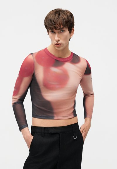 Joven con cabello castaño que lleva una camiseta de manga larga, corta, con un patrón difuminado en rojo y beige, combinada con pantalones negros.