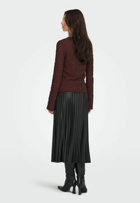 Una donna con lunghi capelli scuri indossa un top bordeaux strutturato, una gonna midi plissettata nera e stivali con tacco neri, in piedi di lato.