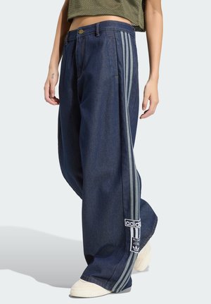 Henkilöllä on yllään tummansiniset leveälahkeiset housut, joissa on kolme harmaata sivuraitaa ja Adidas-logo nilkan lähellä, yhdistettynä valkoisiin lenkkareihin.