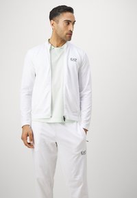 Veste zippée blanche en tissu texturé, assortie à une chemise vert clair et un pantalon blanc coordonné. Avec le logo EA7 sur la poitrine.