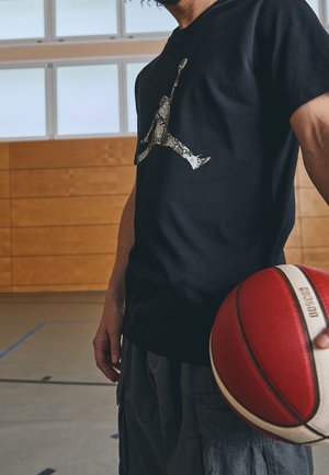 Mand holder en rød og hvid basketball iført en sort t-shirt med en basketballspillerlogo med slangeskindsmønster i et indendørs fitnesscenter.