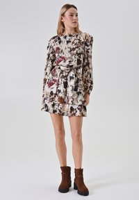 Bloemenjurk met lange pofmouwen, een ceintuur in de taille en een tiered zoom in crème, bordeauxrood en bruine kleuren; gecombineerd met bruine enkellaarsjes.