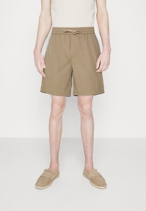 Schlanker Mann, der beige Shorts mit Kordelzug und passende Slip-On-Schuhe trägt, vor schlichtem weißen Hintergrund stehend.