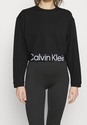 Kvinde iført en sort langærmet crop top med et hvidt 'Calvin Klein'-logo bånd og tætsiddende sorte leggings mod en ensfarvet baggrund.