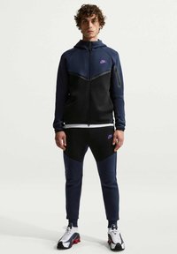 Αθλητική φόρμα σε ναυτικό μπλε και μαύρο ύφασμα, με φούτερ με φερμουάρ, παντελόνι με λεπτή γραμμή και μοβ λογότυπο. Λεπτομέρειες με υφή προσθέτουν ενδιαφέρον.