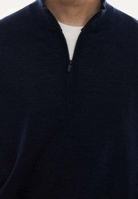 Tmavě modrý svetr s půlvýstřihem na zip vyrobený z texturované látky s límečkem stojačkem a černým zipem. Nasažený přes bílé tričko.