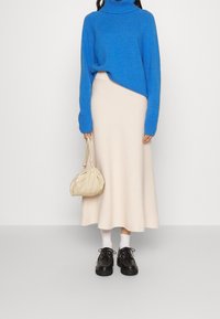 Donna che indossa un maglione a collo alto blu brillante, una gonna midi color crema, calze bianche, scarpe nere e tiene una piccola borsa beige.