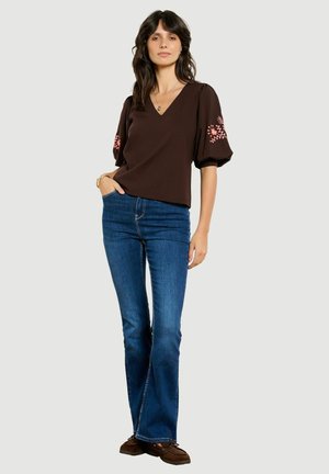 Femme debout, une main dans la poche, portant un chemisier marron foncé à manches bouffantes avec une broderie rose, jean bleu et chaussures marron.