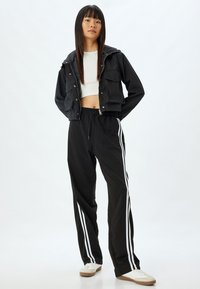 Schwarze, kurze Jacke mit Fronttaschen, kombiniert mit schwarzen Hosen mit weißen Seitenstreifen. Das Model trägt weiße Sneakers.