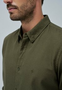 Camicia verde con bottoni e un motivo testurizzato, dotata di colletto e di un piccolo logo sul petto, mostrata sul profilo laterale di un modello.