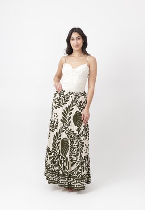 Mujer que lleva una blusa corsé sin mangas blanca y una falda larga color crema con grandes motivos de hojas verde oscuro, de pie frente a un fondo liso.